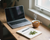 Laptop on a wooden desk with a notebook, pen, and steaming cup of tea near a window.